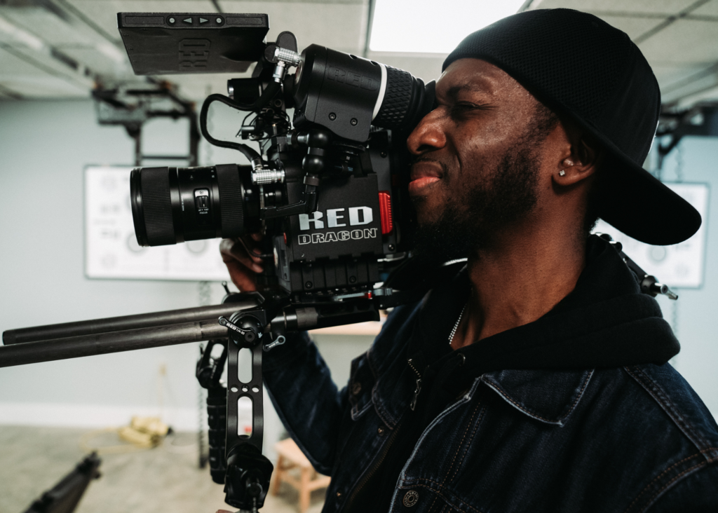 A Black man in a denim jacket, hoodie, and black baseball cap stands holding a film camera that points to the left of the frame. He is looking through a viewfinder, and the eye facing the camera is closed.