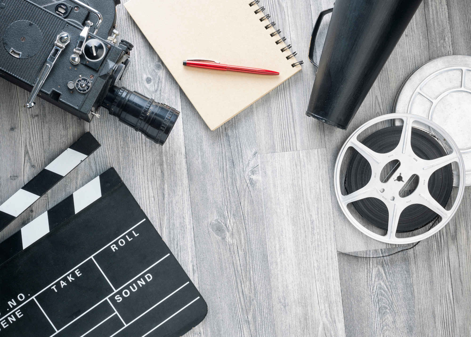 A flat lay of film equipment includes a clapboard, a vintage camera, a notebook with a red ballpoint pen, the speaking end of a vintage film megaphone, and a reel of film - all lying on a light gray wood grain background.