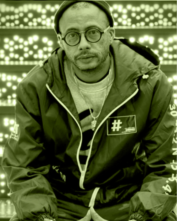 A green tinted photo shows a Black man in a windbreaker, beanie and glasses leaned forward toward the camera. His background is pixelated, almost as if lit by a Lite-Brite.