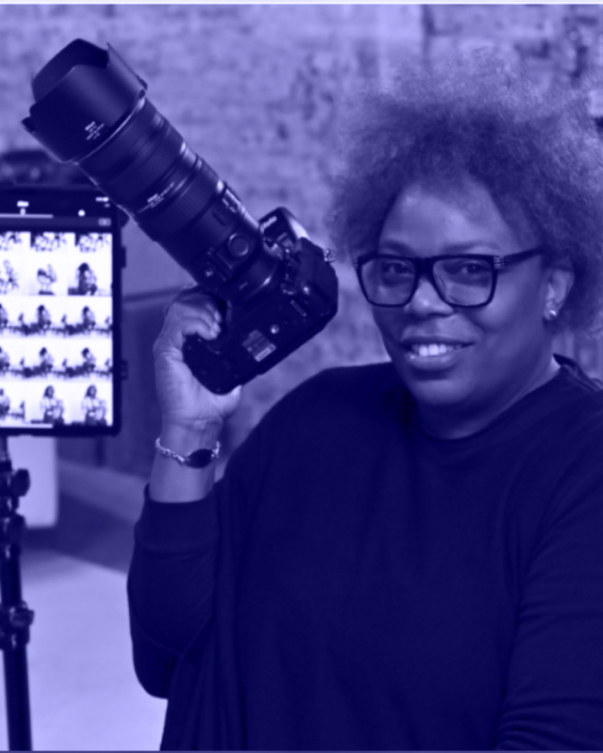 A blue tinted photo shows a Black woman in glasses and with Natural hair holding up an SLR camera with a lens on. To her right is a monitor showing thumbnails of her captured photos.