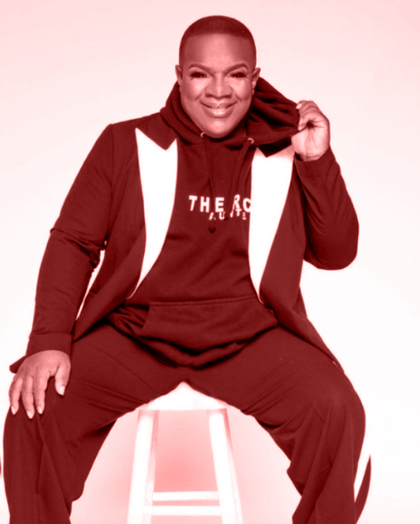 A pink tinted photo has a Black person sitting on a stool in a sweatsuit and a blazer, holding the bottom edge of the hoodie out and smiling.