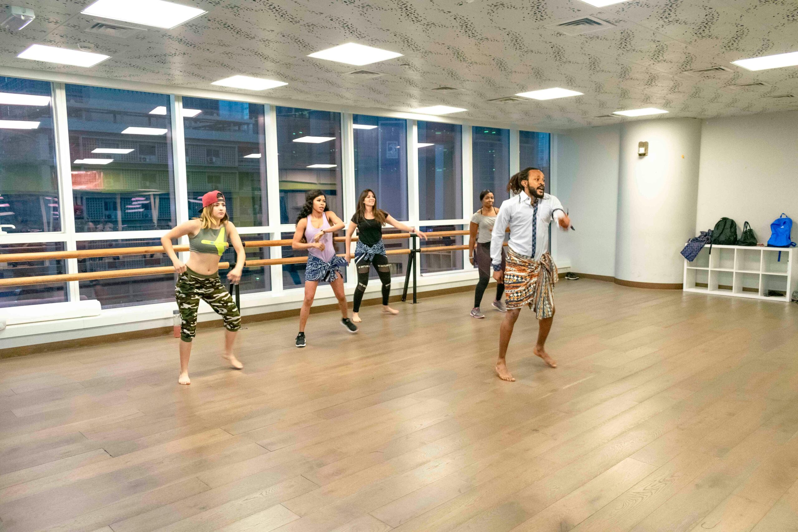 In a dance studio with a window as its back wall, a young man leads a group of young women in a dance routine. He demonstrates a move as the women stand back and watch his progress.