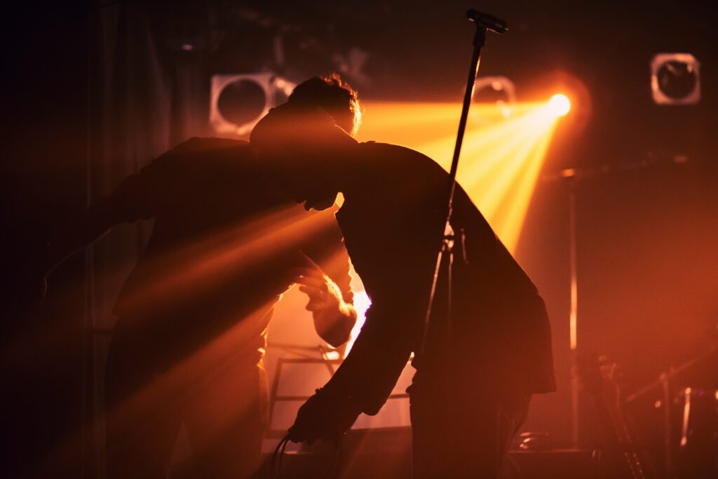 Lit warmly by a orangish light shining from the background, two people work to warangle cords in front of a mic stand.